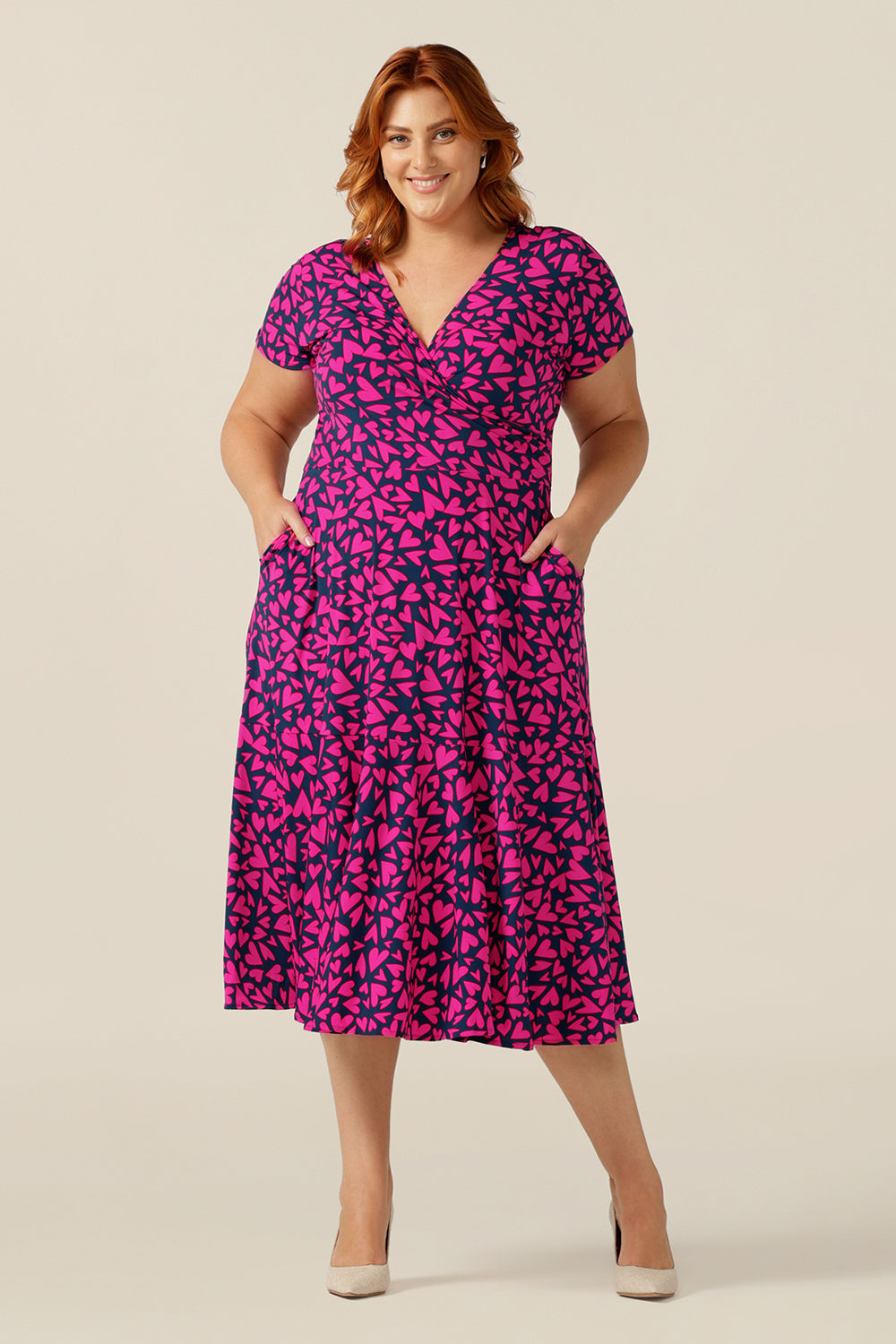 size 18 curvy woman wears a reversible jersey wrap dress. Worn with the crossover neckline to the front, this dress can be worn reversed with a boat neckline. This dress has short sleeves and a below-the-knee skirt with pockets. The dress is patterned with a pink heart print - perfect as a Valentines Day date-night dress or evening or occasionwear.