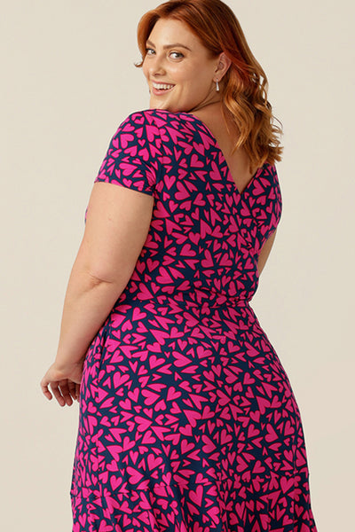 size 18 curvy woman wears a reversible jersey wrap dress. Worn with the crossover neckline to the front, this dress can be worn reversed with a boat neckline. This dress has short sleeves and a below-the-knee skirt with pockets. The dress is patterned with a pink heart print - perfect as a Valentines Day date-night dress or evening or occasionwear.