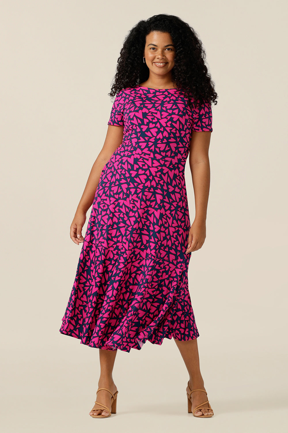 a size 12 curvy woman wears a reversible jersey wrap dress. Worn with the boat neckline to the front, this dress can be worn reversed with a crossover, V-neck. This dress has short sleeves and a below-the-knee skirt with pockets. The dress is patterned with a pink heart print - perfect as a Valentines Day date-night dress or evening or occasionwear.