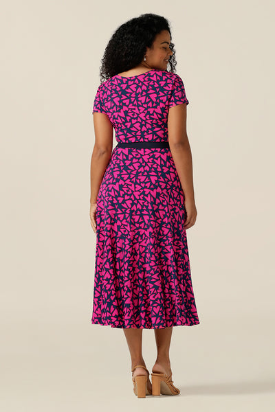 a size 12 curvy woman wears a reversible jersey wrap dress. Worn with the boat neckline to the front, this dress can be worn reversed with a crossover, V-neck. This dress has short sleeves and a below-the-knee skirt with pockets. The dress is patterned with a pink heart print - perfect as a Valentines Day date-night dress or evening or occasionwear.