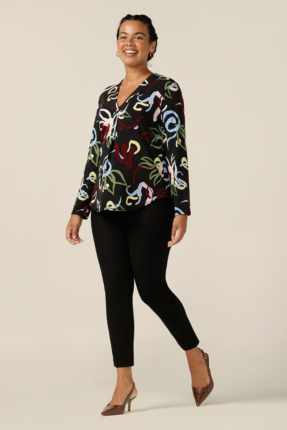 A size 12, curvy woman wears a V-neck jersey top with long sleeves and a shirttail hemline. Australian-made by Australia and New Zealand women's clothing label, L&F, this long sleeve top features an abstract print with a black base. This workwear top is worn with black trousers for easy corporate style.