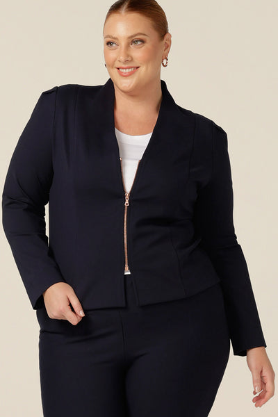 A size 18, fuller figure women wears a corporate navy jacket. A soft tailored jacket with rose gold zip fastening, long sleeves and a collarless neckline, this workwear jacket is worn with navy tailored trousers for a corporate look.