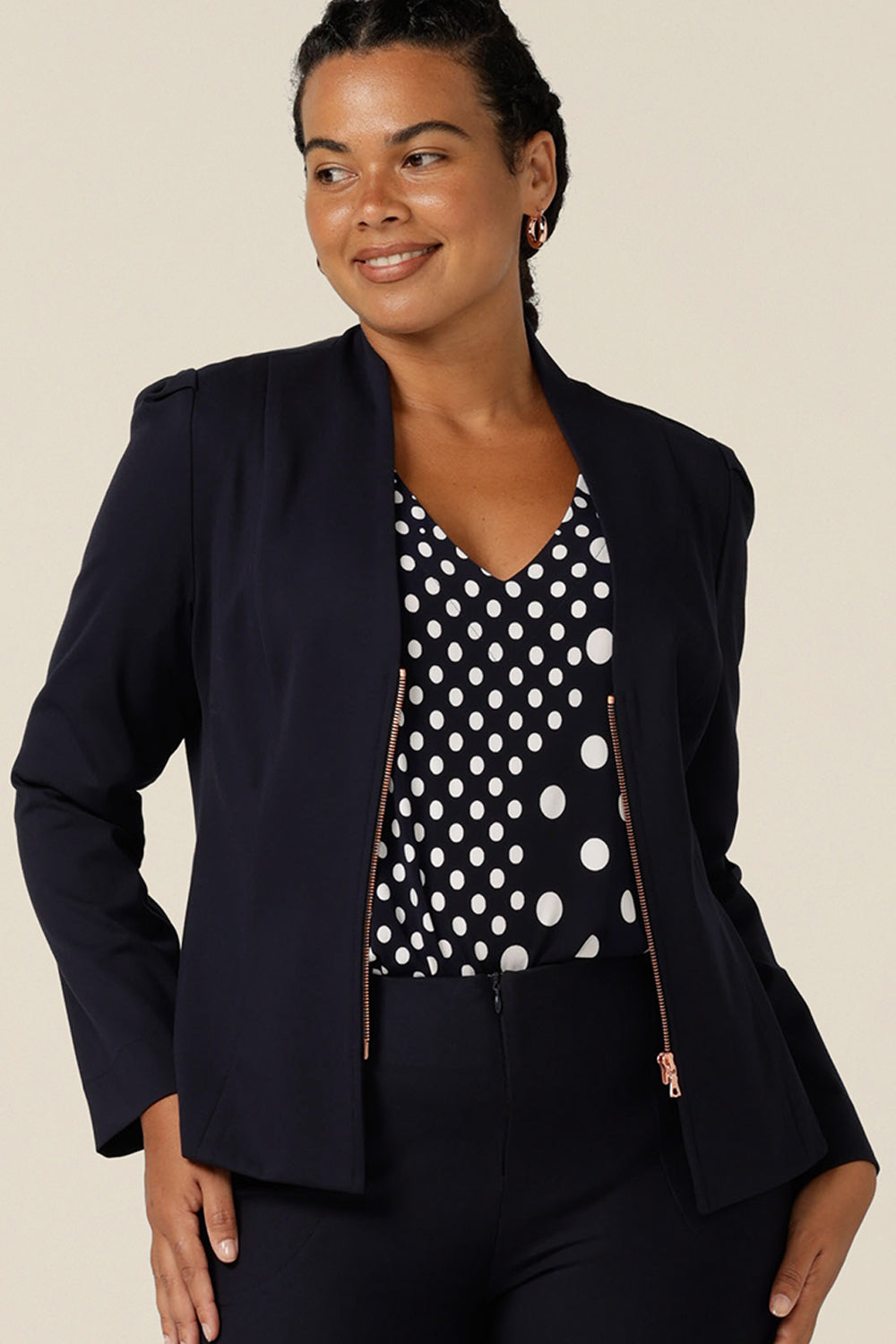 A size 12, curvy woman wears a corporate style, navy jacket with navy and white jersey top. A soft tailored jacket with rose gold zip fastening, long sleeves and a collarless neckline, this workwear jacket is worn with navy tailored trousers.