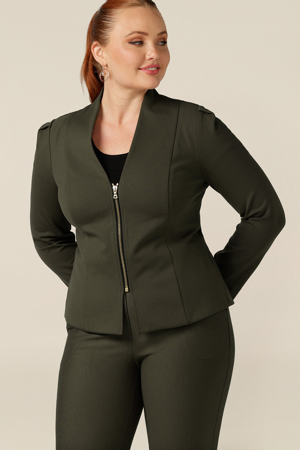 A size 12, curvy woman wears a tailored jacket with collarless V-neckline and zip fastening. Australian-made in olive green ponte jersey, the stretch fabric makes for a comfortable corporate jacket.