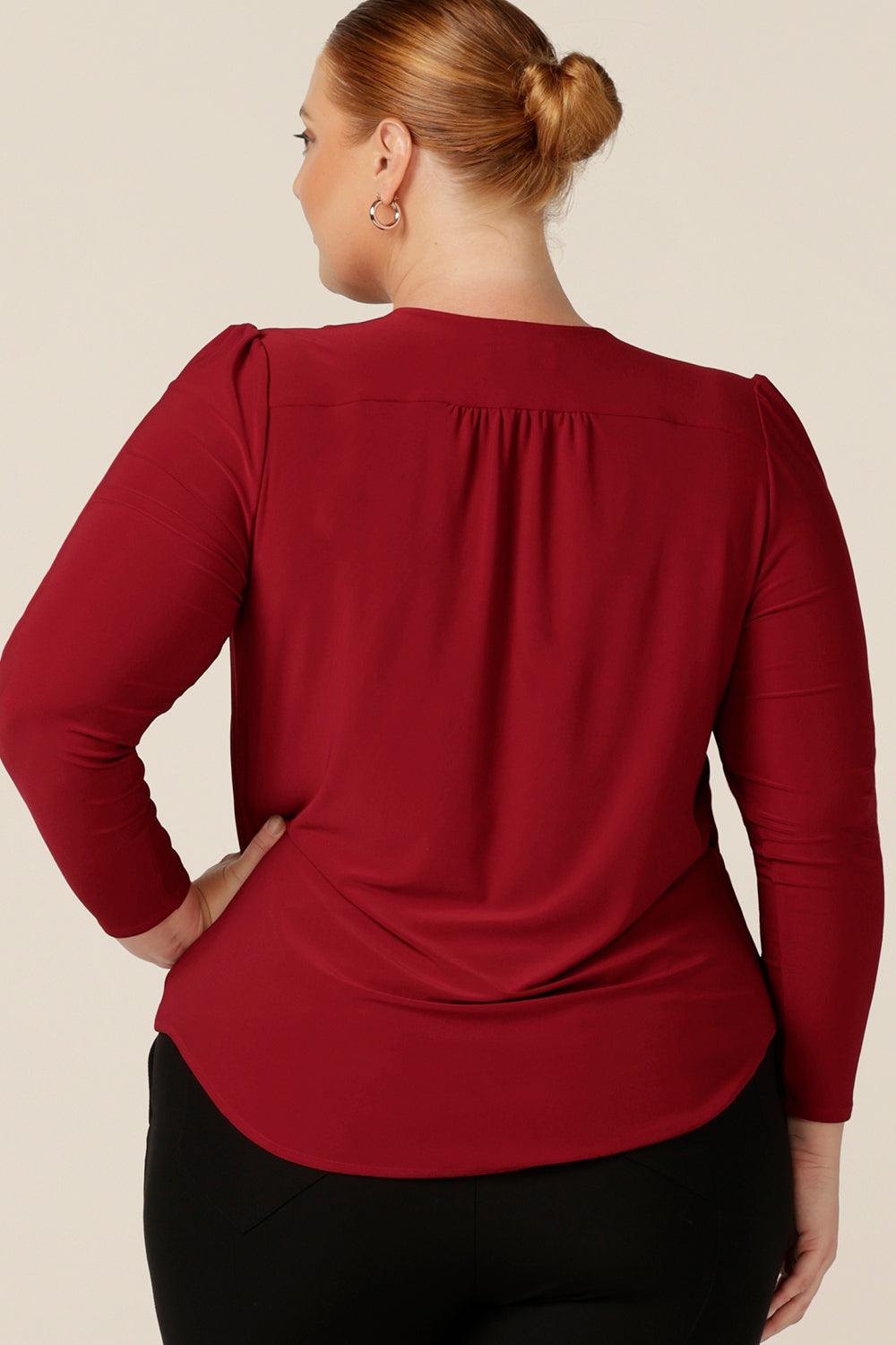 Back view of a classic long sleeve women's top in flame red jersey. The perfect top for workwear and casual style, this Australian-made top features a V neck, shirttail hemline and long sleeves. Worn by a fuller figure, size 18 woman, this jersey top comes in an inclusive size range of sizes 8-24. 