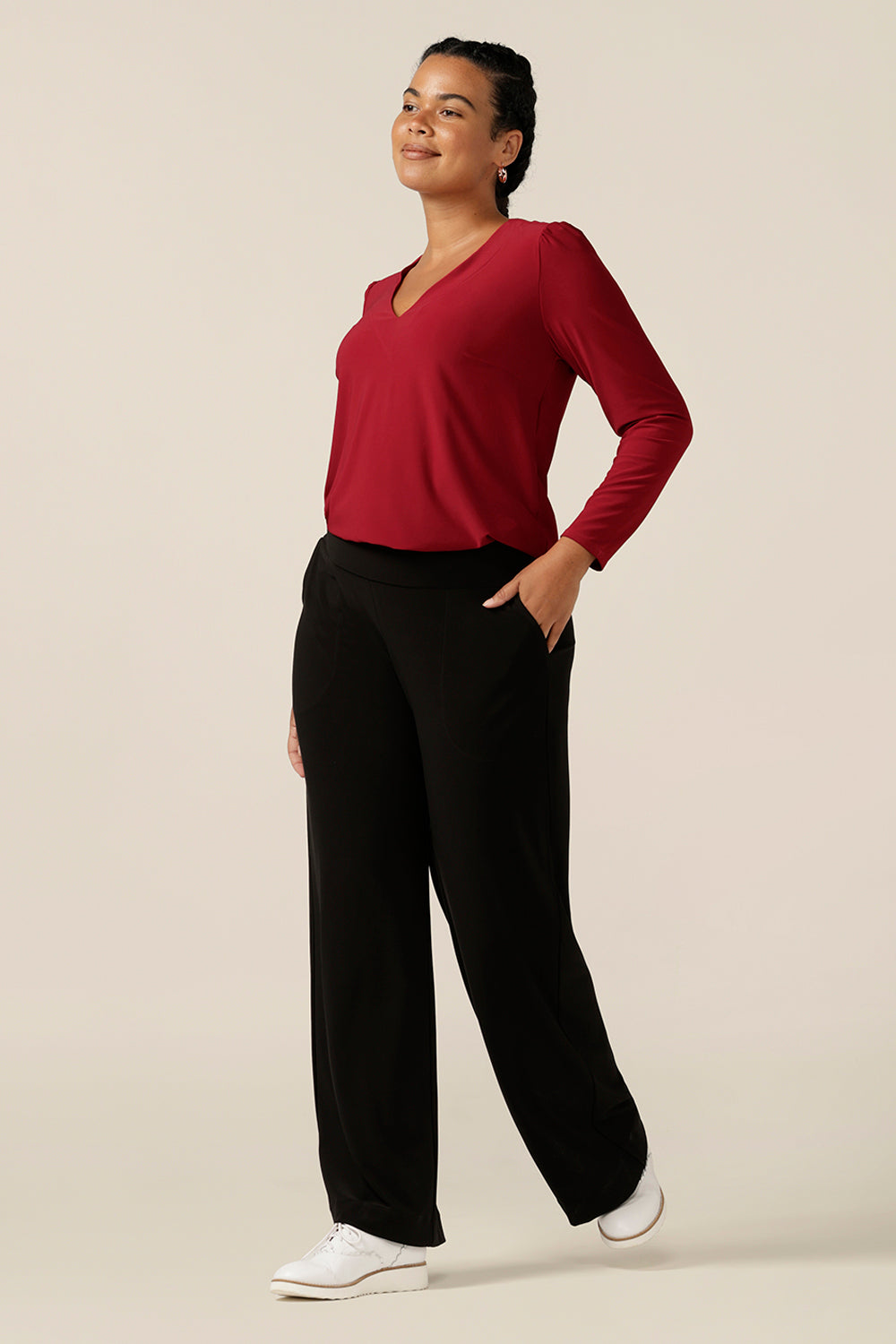 A size 12, curvy woman wears a V neck jersey top in flame red jersey. With long sleeves, this top is a good autumn/winter top, and is styled here with wide leg black pants for a casual look. 