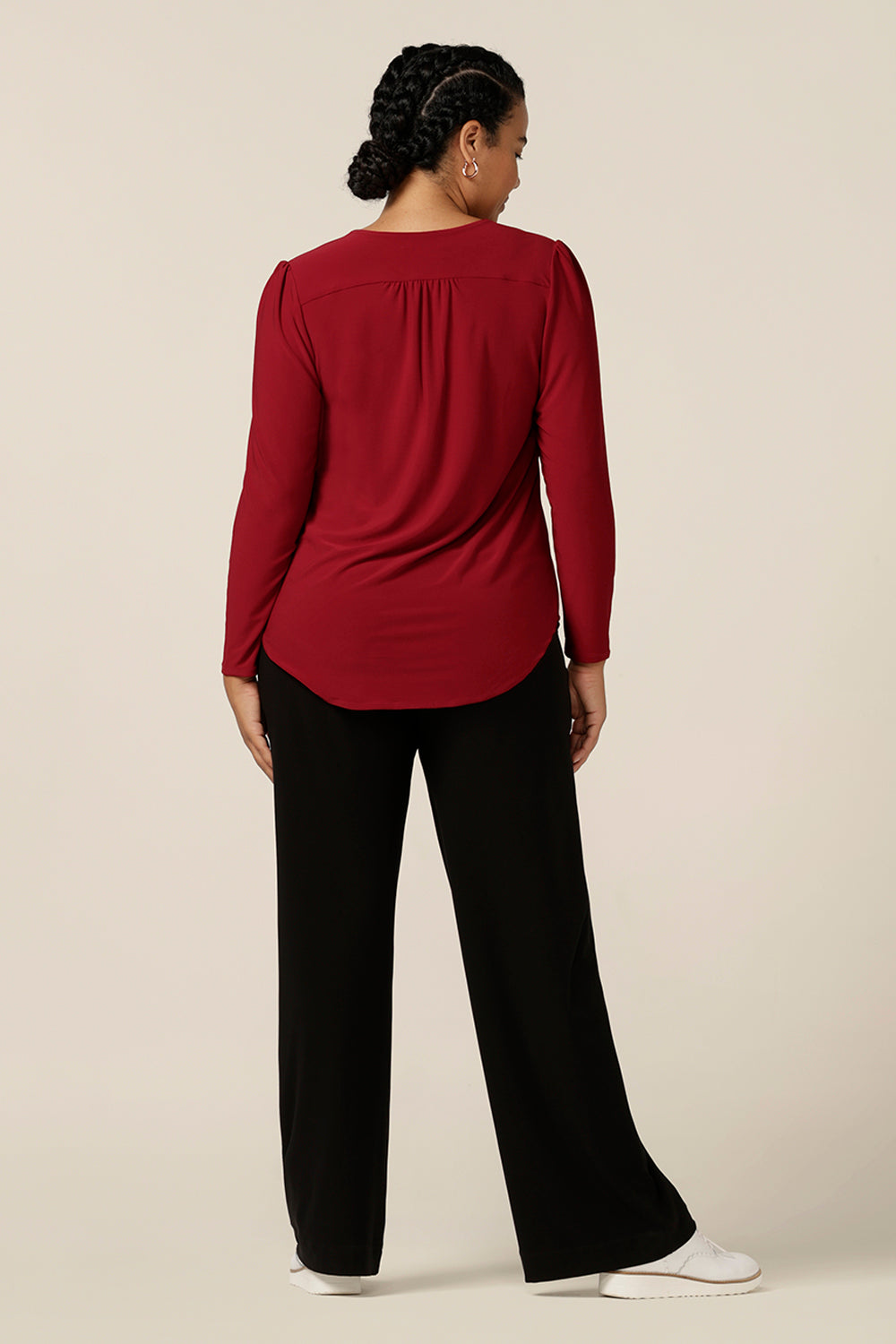 Back view of a size 12, curvy woman wearing a V neck jersey top in flame red jersey. With long sleeves, this top is a good autumn/winter top, and is styled here with wide leg black pants for a casual look.
