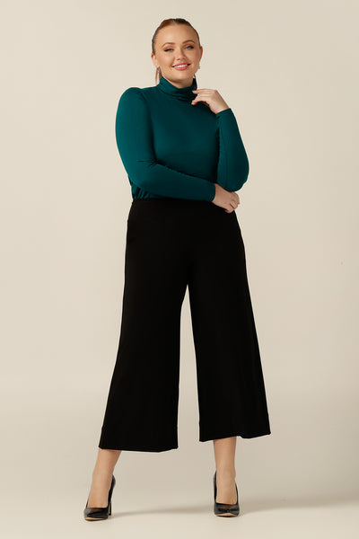 Back view of an Australian-made, women's long-sleeve, turtle neck top in petrol green bamboo jersey. Worn with wide leg, cropped length black workwear pants for a corporate outfit.