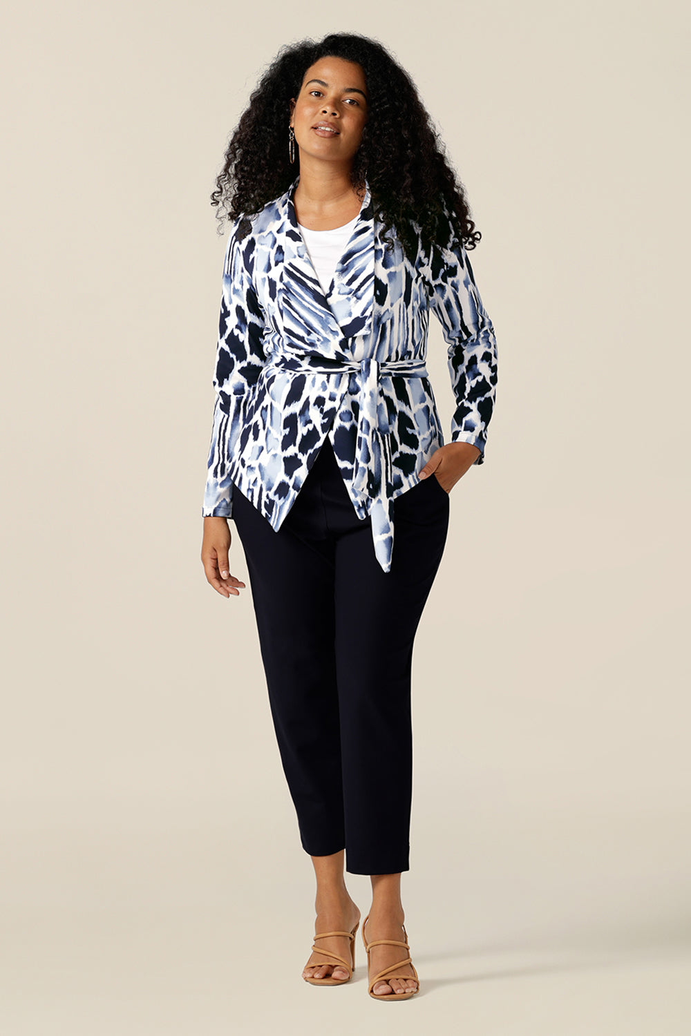 A size 12 curvy woman wears a wrap-over jacket in comfortable stretch jersey. Patterned in navy blue and white, this jacket has a V-neck, long sleeves and a belt tie. Designed as a work wear jacket, the Hayden Jacket is worn with navy tapered leg cropped pants for a work wear outfit. Made in Australia, quality office-wear jackets come are ready to shop in sizes 8 to size 24 at Leina and Fleur.