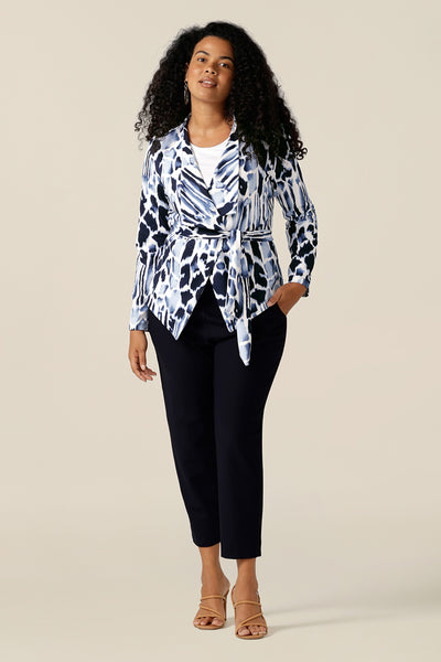 A size 12 curvy woman wears a wrap-over jacket in comfortable stretch jersey. Patterned in navy blue and white, this jacket has a V-neck, long sleeves and a belt tie. Designed as a work wear jacket, the Hayden Jacket is worn with navy tapered leg cropped pants for a work wear outfit. Made in Australia, quality office-wear jackets come are ready to shop in sizes 8 to size 24 at Leina and Fleur.
