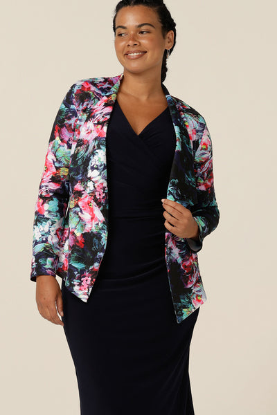 A size 12, curvy woman wears a wrap around jacket in comfortable stretch jersey. Patterned in an abstract floral print on a black base, this jacket has a V-neck, long sleeves and a belt tie. Designed and made in Australia, shop workwear jackets in sizes 8-24 now with free shipping to New Zealand.