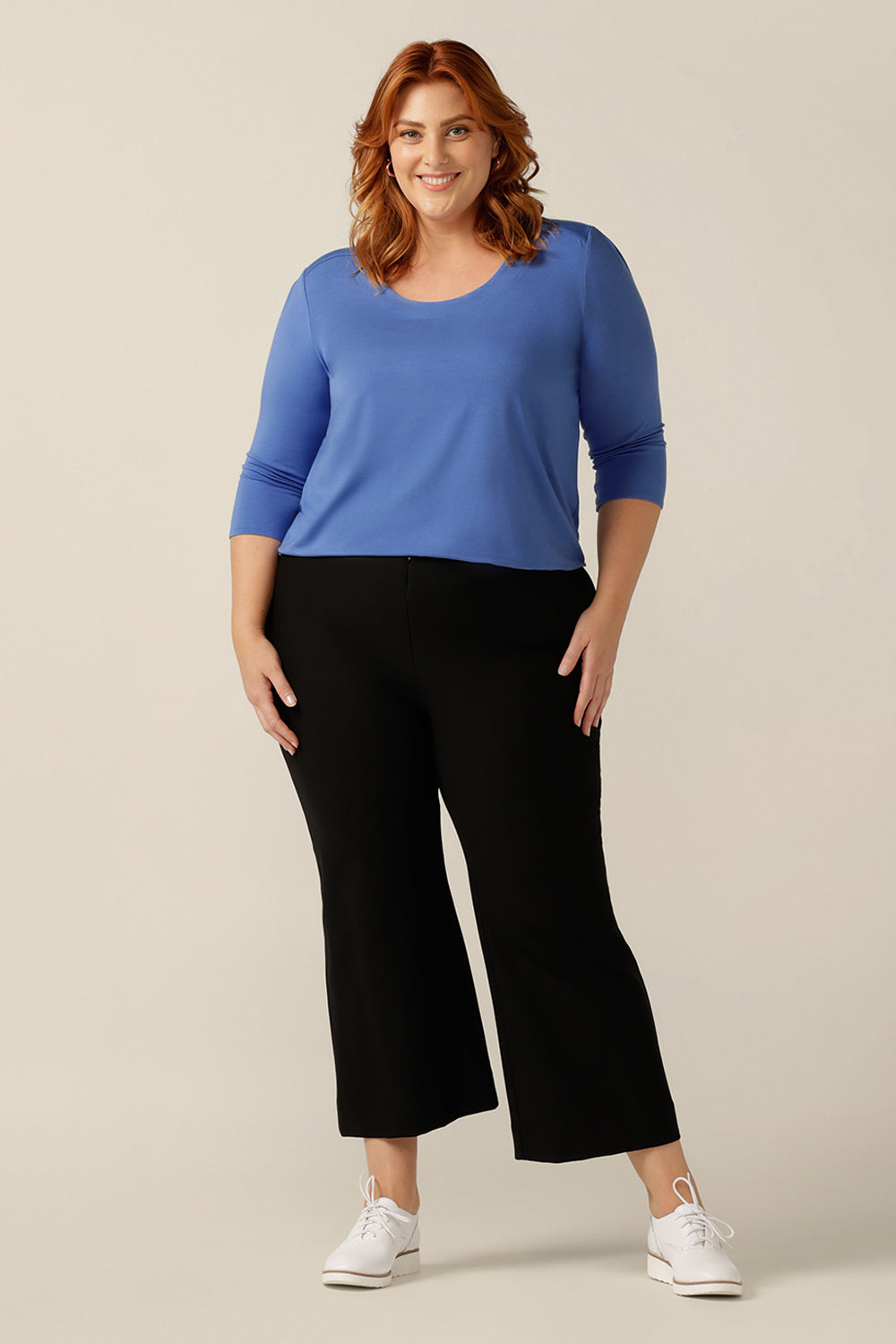A plus size, size 18 woman wears a blue bamboo jersey top. The top has a round neckline, 3/4 sleeves, and a curved shirttail hemline. Worn with black work wear pants, tuck in this bamboo jersey top and she becomes a smart-casual work wear top. 
