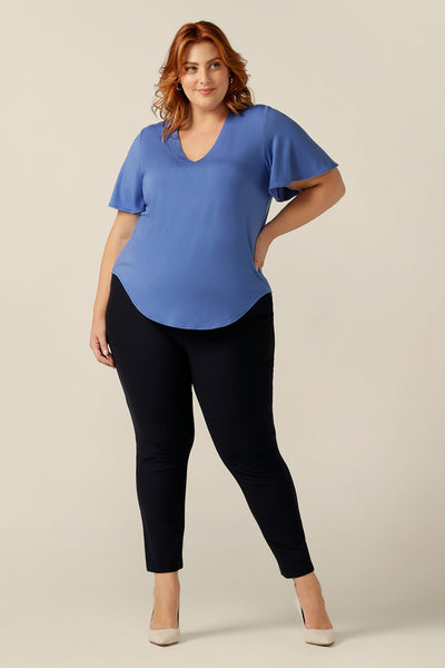 A plus size, size 18 woman wears a V-neck blue top with short flutter sleeves. Made from bamboo jersey, this is a natural fibre top that is lightweight, breathable and sustainable. The blue bamboo jersey top is worn over slim-leg black jersey trousers for a smart-casual look.