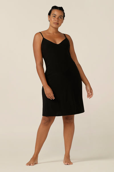 A size 12 woman wears a mini-length, reversible slip in black, slinky jersey fabric. The slip has thin straps and is worn with a V neck to the front. Made in Australia to layer under dresses and separates. Plus sized lounge wear plus size slip dress