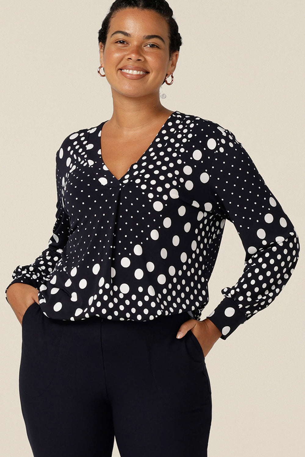 A size 12 curvy woman wears a long sleeve, V-neck top in navy and white polka dot print jersey. Australian-made, this top is good for workwear style. 