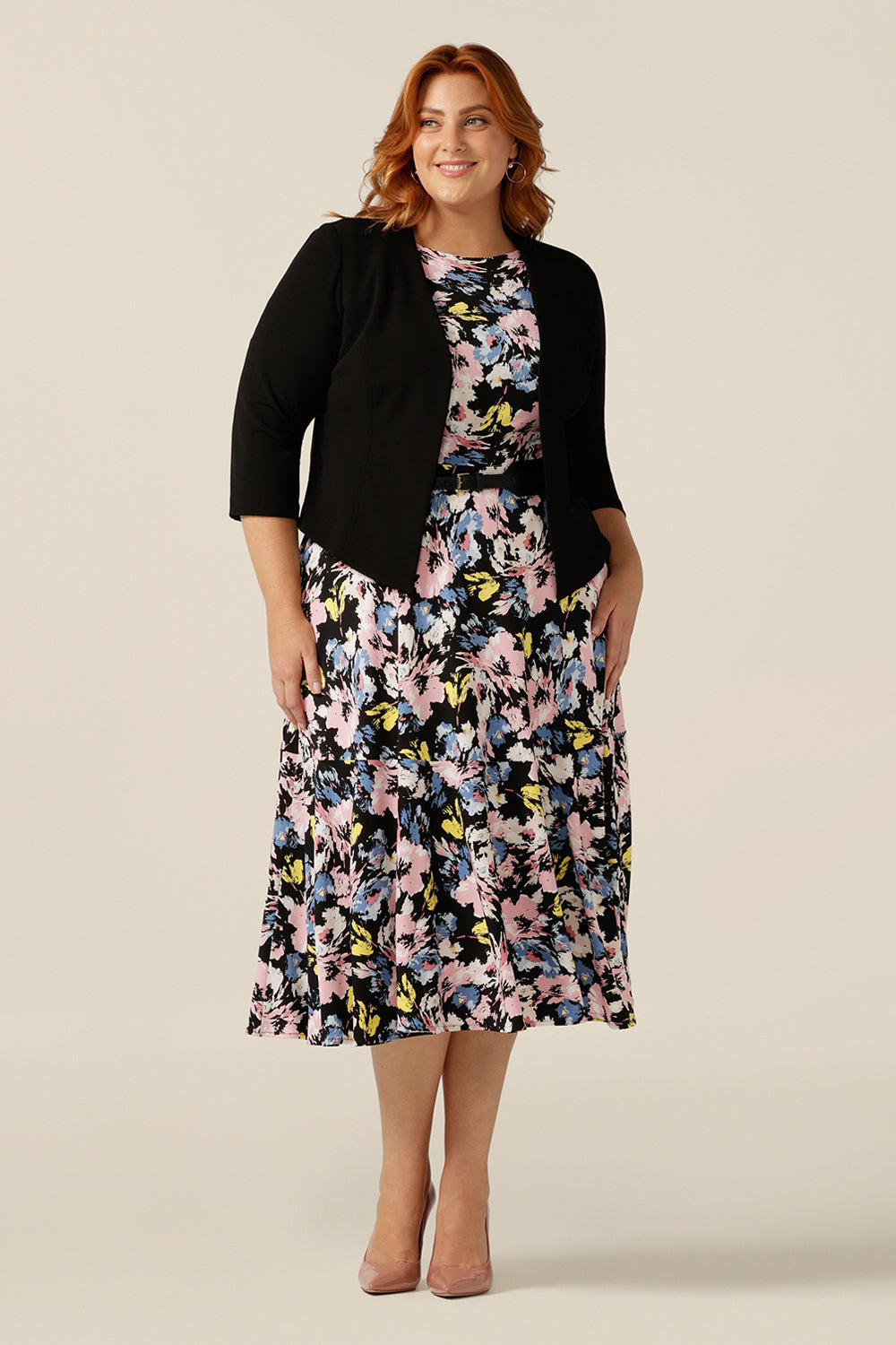 A plus size, size 18 woman wears a black workwear jacket over a floral print jersey dress. The Rainy Jacket is a collarless, open front jacket with cropped sleeves. Made in crepe jersey, this is a soft tailoring jacket with stretch for a comfortable twist on quality-made work wear suit jackets for women.  