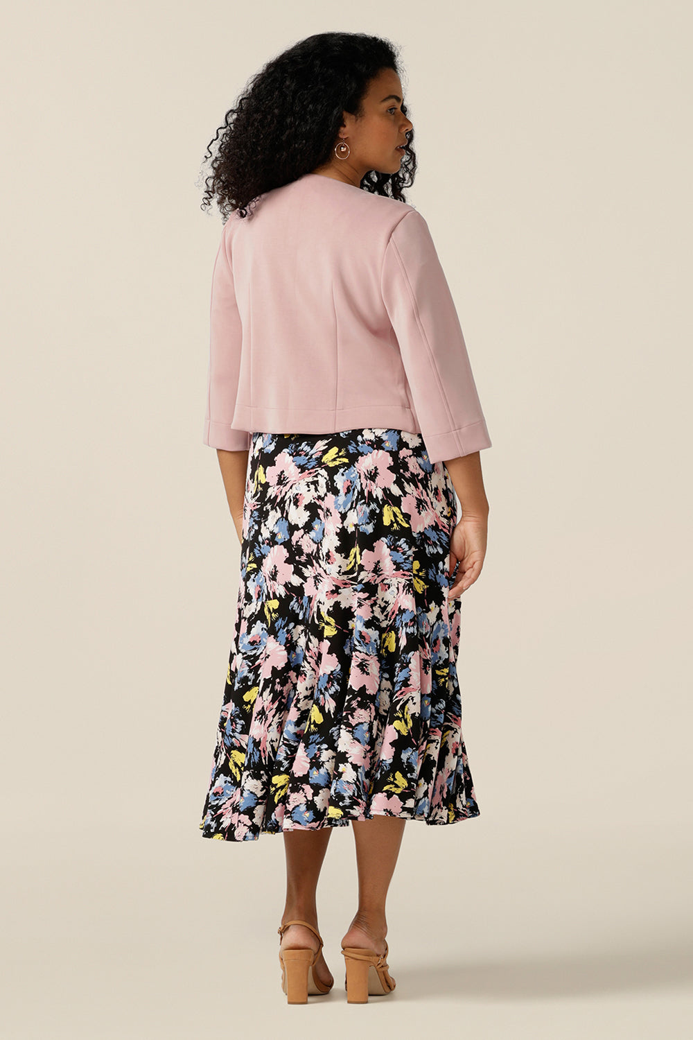 A size 12 curvy woman wears an open-front, collarless jacket with 3/4 sleeves. Made from pink modal jersey, this comfortable jacket is sustainably-made using natural fibres and eco-conscious production. A good sustainable corporate jacket, the Rainy Jacket is worn with a vintage floral print jersey dress.
