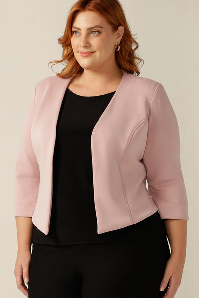 a plus size, size 18 woman wears a soft tailoring collarless jacket in pink modal. A fitted jacket for work, the stretch fit of the modal jersey makes for a comfortable corporate jacket. Worn with a black top and black trousers to give a sleek smart casual work outfit.