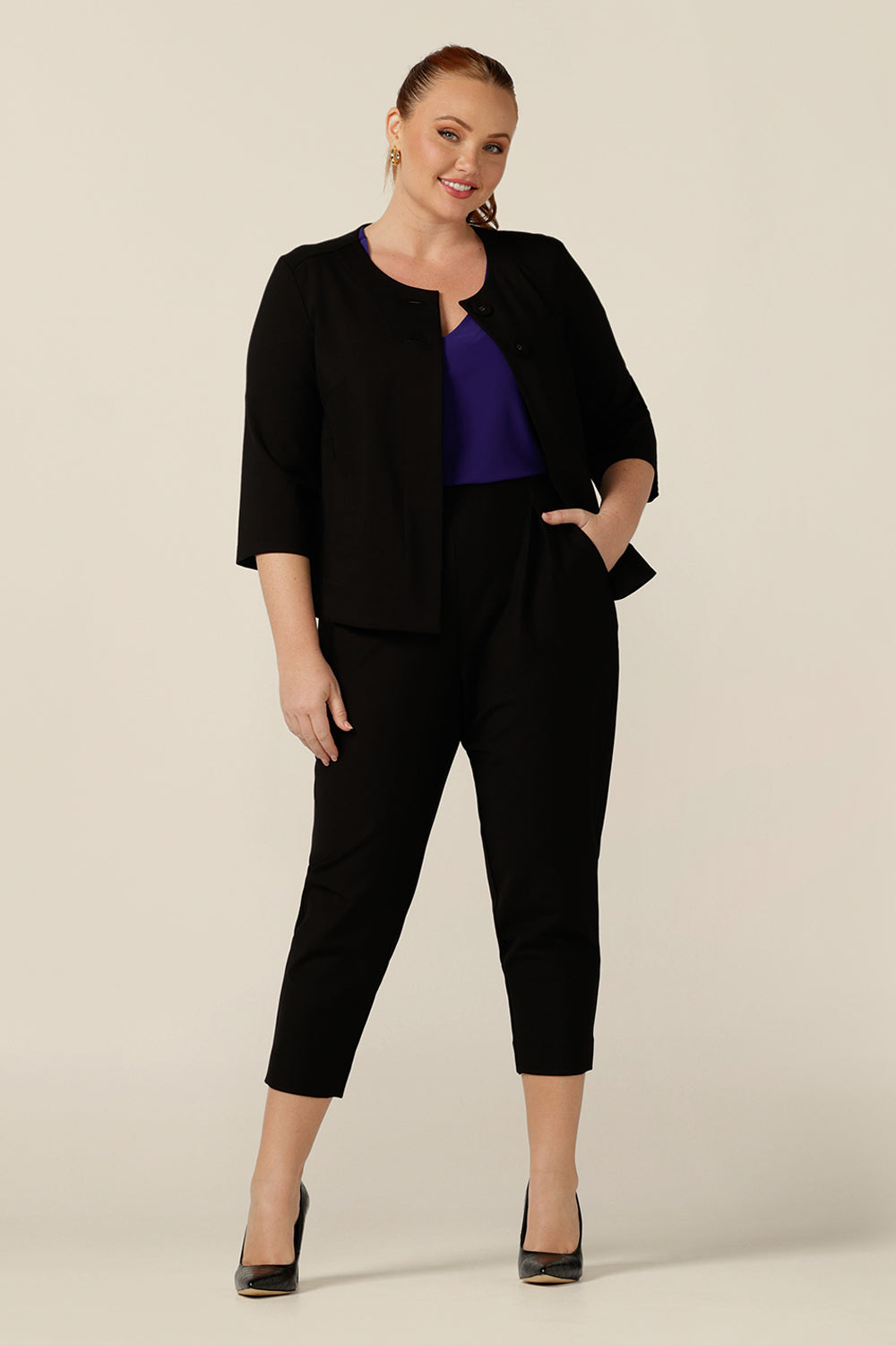A size 12 woman wears a collarless black jacket with 3/4 sleeves over a purple jersey top and tapered-leg cropped black pants to create a work wear outfit. Cut for corporate comfort, this work jacket swings to the back and fits petite to plus size women.