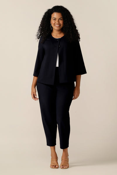 A curvy size 12 woman wears a round neck navy work wear jacket with 3/4 sleeves and a swing back.