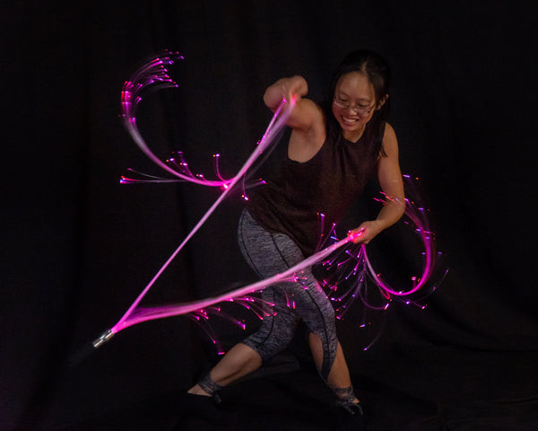 A fiber whip dancer taking a dramatic lunge pose with a split whip, forming glowing pink trails of light behind her.