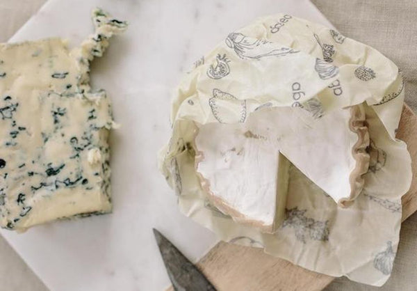 Abeego beeswax wrap around a wheel of soft brie cheese atop a wood cutting board and next to a bright wedge of blue cheese 