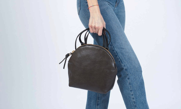 Woman wearing jeans holding a black Eleven Thirty Anni bag with a circular top, flat bottom, and single top handle
