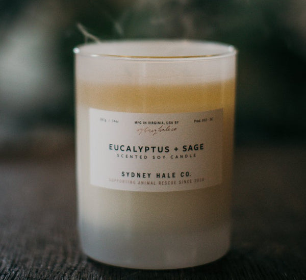 Sydney Hale eucalyptus and sage soy candle in a frosted glass container sitting on a wood table with soft plumes of smoke 