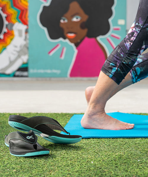 a woman takes off her open heel sandals to perform yoga at the park.