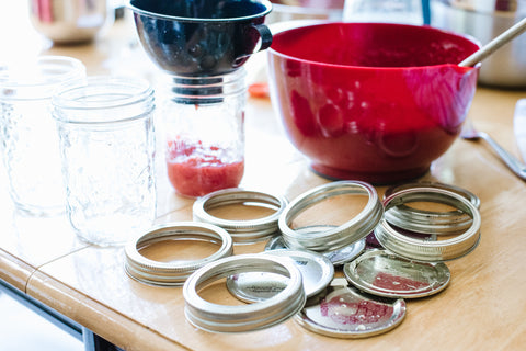 wooden table covered with mason jars and lids and a red bowl full of strawberry freezer jam