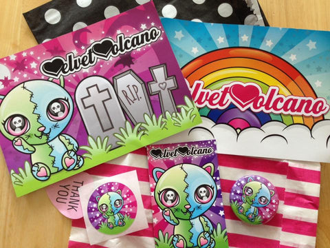 Photo shows 2 flyers, 2 small paper bags, a sticker, a button badge and a business card on a wooden backdrop. To the left of the image the first flyer features a cartoon cat inspired by Frankenksteins Monster in a graveyard with tombstones and bats, the sky is purple. The flyer to the right of this features a rainbow with clouds underneath it and a sunburst style turquoise background with stars. Above the flyers is a black paper bag with white polka dots, laying flat. Below the flyers is a bright pink and white striped paper bag, laying flat. On top of this bag is a round sticker featuring the FrankenKitty design, next to a business card with the same design and on the right of that is a FrankenKitty badge design.