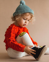 Baby sitting down wearing a blue beanie, red sweater, smiley leggings and charcoal Glerups wool baby boots