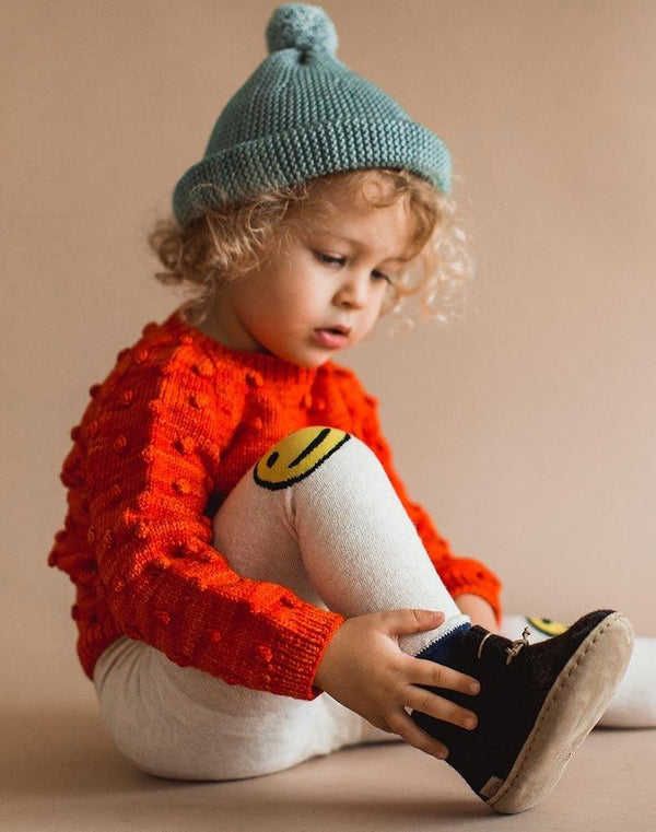 Baby sitting down wearing a blue beanie, red sweater, smiley leggings and charcoal Glerups wool baby boots