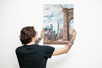A hipster young man with tattoos holding a 'Create Your Own' metal poster display plate of the Brooklyn Bridge in New York 