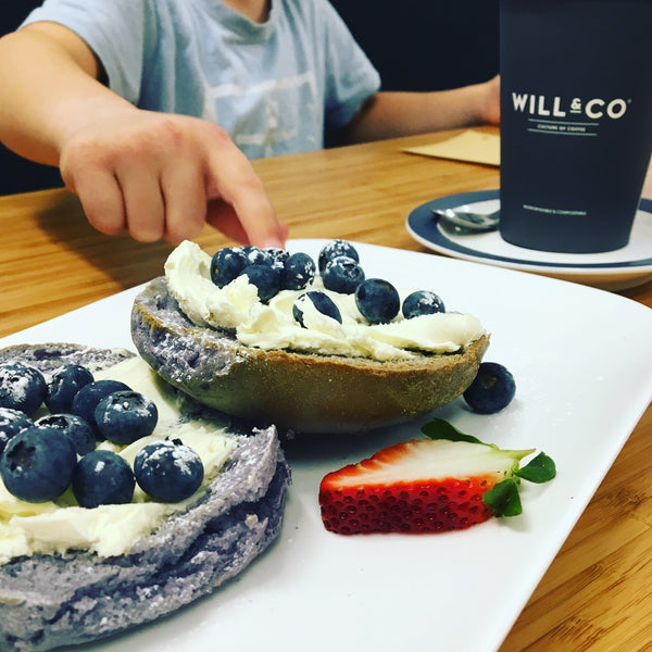 Blueberry bagels in the foreground with a child's hand stealing blueberries off the plate