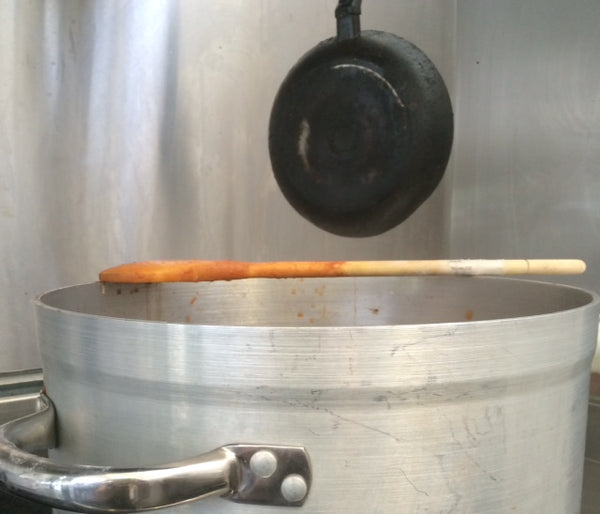 Silver pot on the stove, with a wooden spoon resting on the rim, below a black hanging ladle