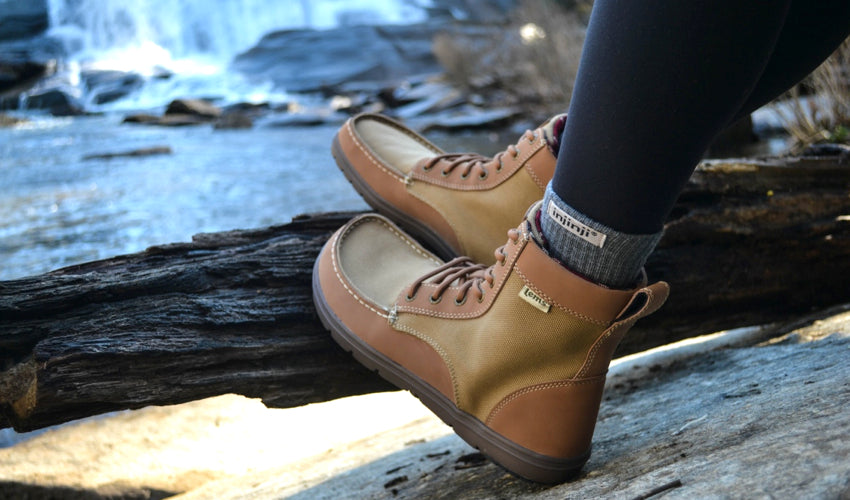 Person resting their Lems Boulder Boot Brown boots on a log with a waterfall and stream in the background