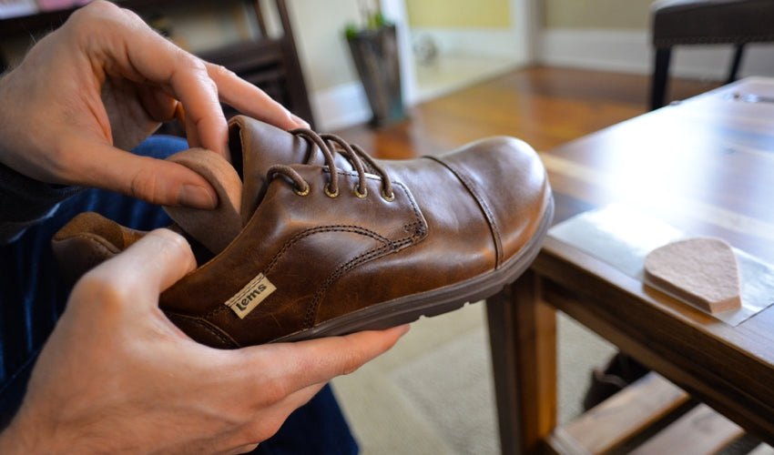 A person placing a tongue pad within a pair of Lems Nine2five Mocha shoes