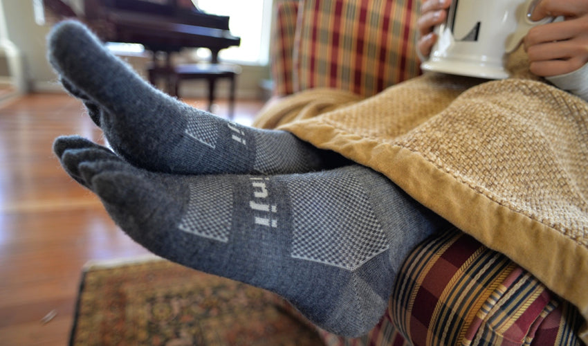 The lower legs of a person seated beneath a blanket on a couch wearing Injinji Original Weight Crew Wool Charcoal toe socks