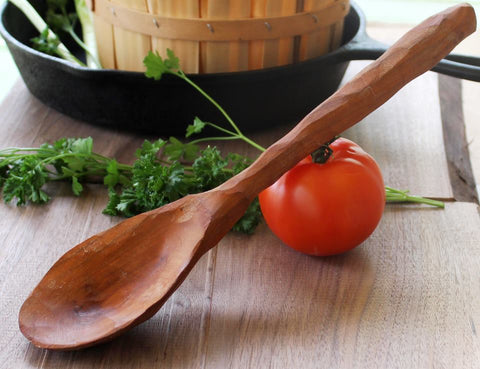 Handmade wooden cherry spoon leaning on a tomato with herbs on a brown background with a black pot
