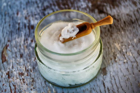 Jar of coconut oil with wood scoop containing a dollop against an off-white background