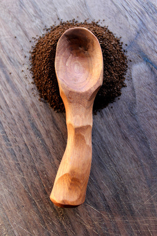 Handmade wooden coffee scoop against a background of ground coffee and a brown tabletop.