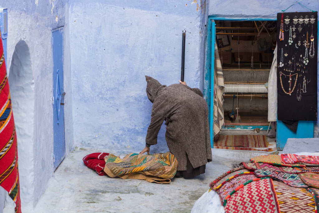 Street Scenes in Chefchaouen, Morocco by Sophee Smiles - Hooded Man Bending Over Rugs
