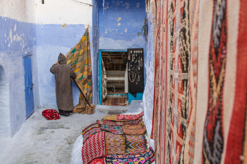 Street Scenes in Chefchaouen, Morocco by Sophee Smiles - Hooded Man Hanging up Rugs