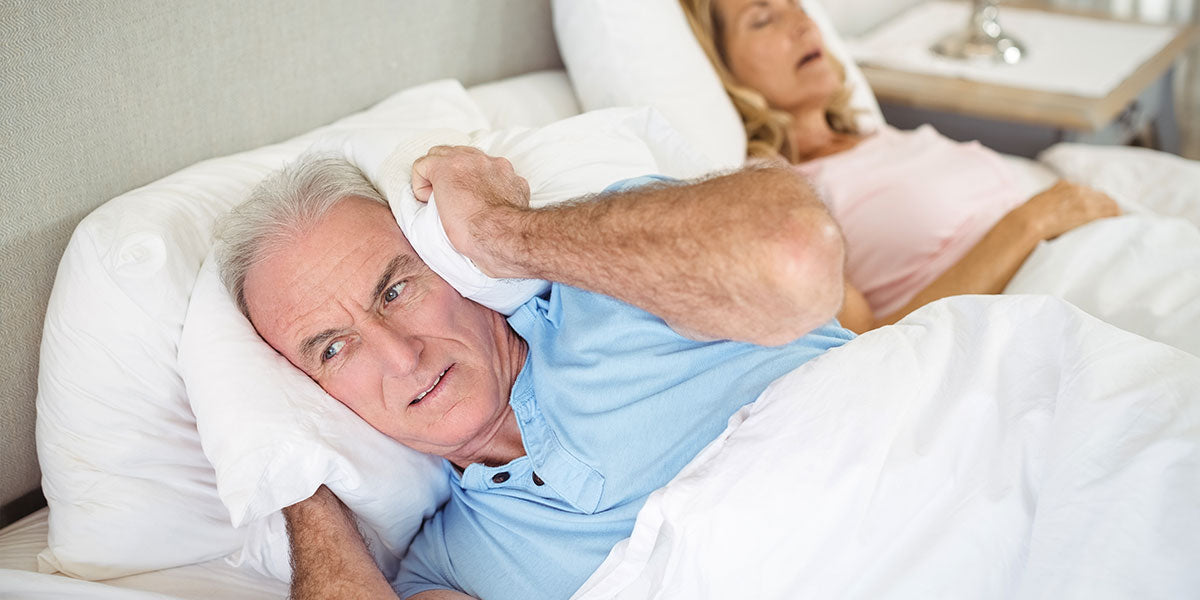 Senior man lying on bed and covering his ears with pillow in bedroom
