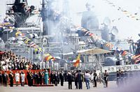 Smoke clears around the guided missile cruiser USS BELKNAP (CG 26) and other ships following a gun salute in honor of Spain's King Juan Carlos and Queen Sophia, pictured on the pier, at center. The royal couple is in Barcelona to preside over an international naval review which includes the BELKNAP and the Spanish navy's new aircraft carrier. - 1990