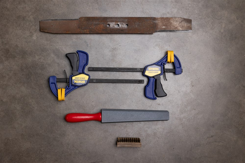 The tools needed for sharpening a lawn mower blade, the removed blade, clamps, a wire brush and an appropriate sharpener.