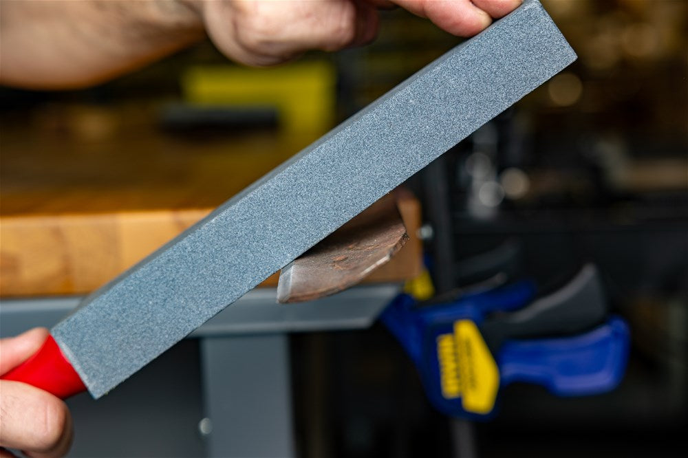 The Norton Utility File is held against the lawn mower blade bevel to match the existing sharpening angle.