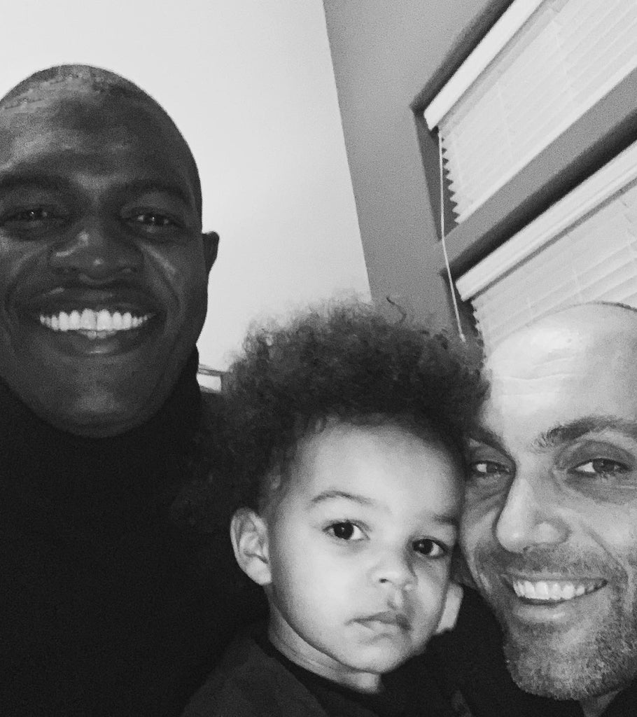 Black and white photo of smiling fathers, Kwesi and Jackson, with their young son, taking a selfie at home. 