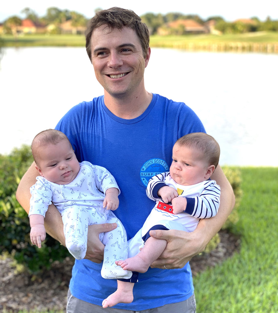 A gay dad holds both of his twin babies in his arms, standing in a grassy park in front of a lake.